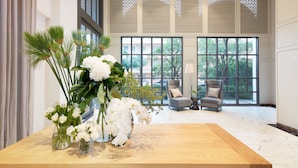 Een ruime kamer met grote ramen, een houten tafel met een vaas witte bloemen en twee grijze fauteuils.