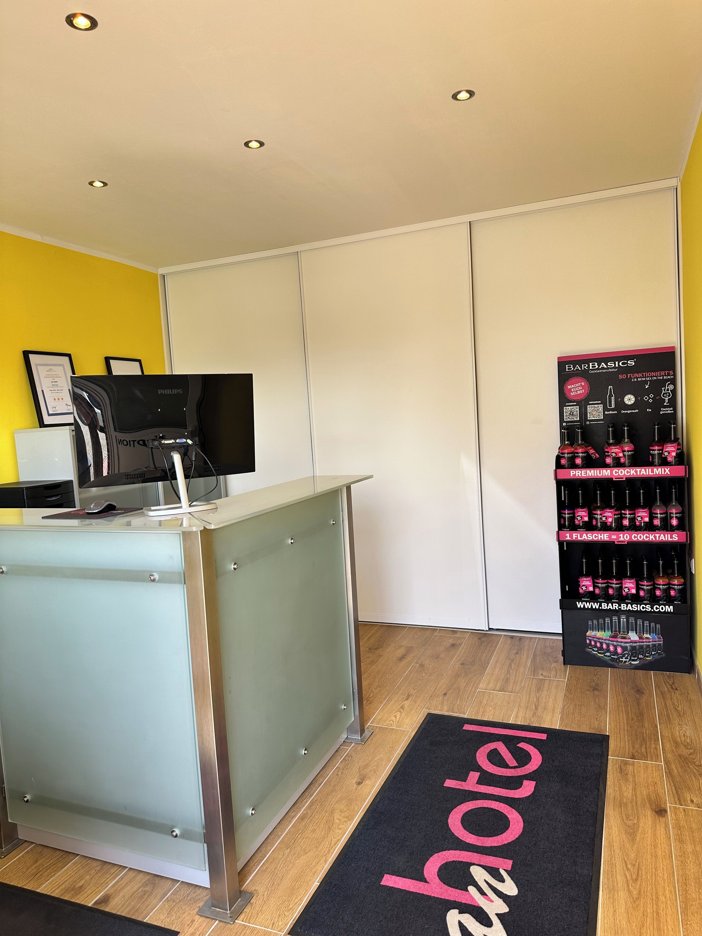 A reception desk with a computer and a flat-screen TV, a display of Barbasics cocktail mix, and a doormat with "hotel" written on it.
