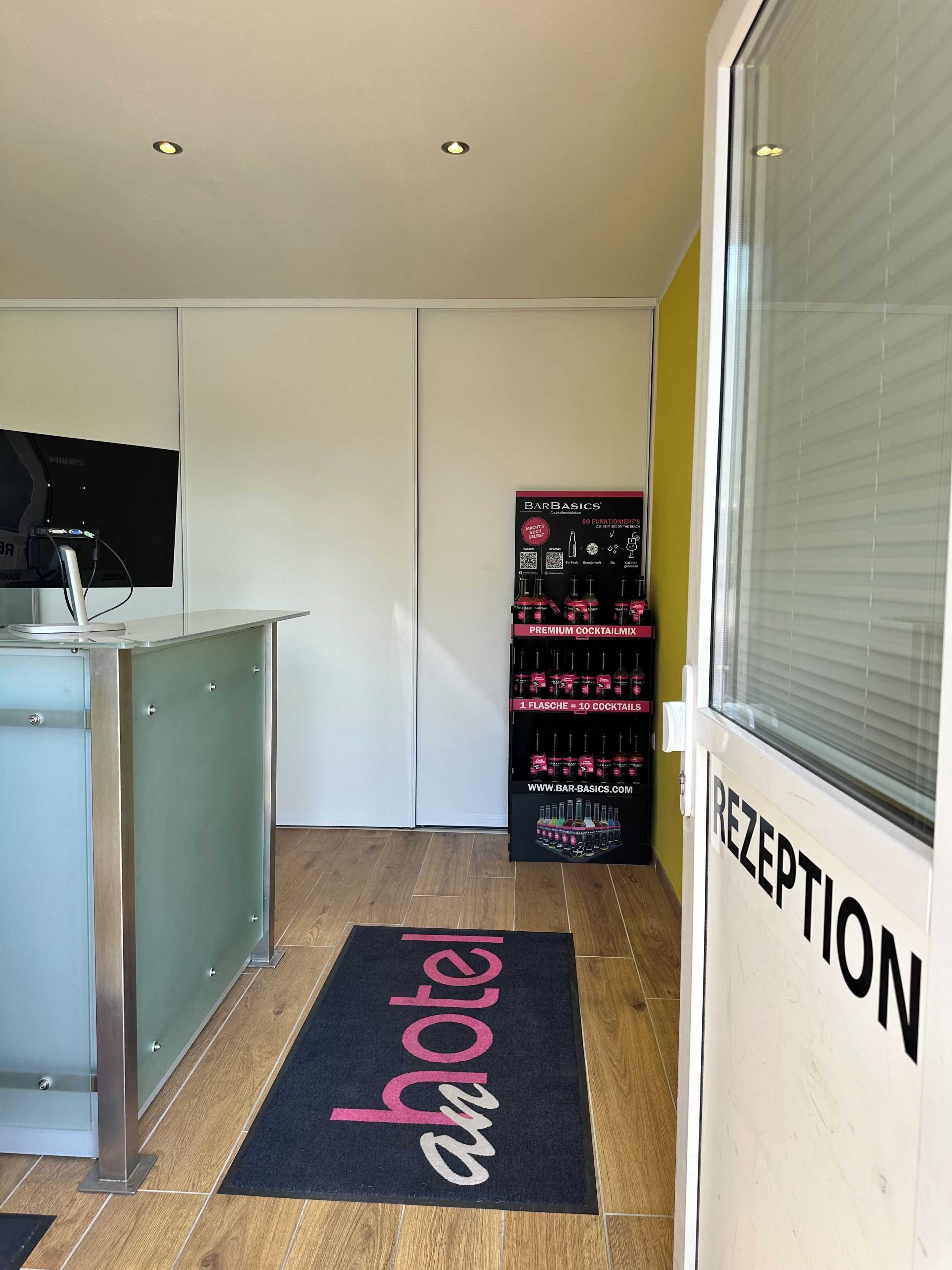 En hotellobby med en receptionsdisk, en digital skärm, en receptionsskylt, en matta med texten ”hotell” och en hylla med flaskvatten och andra artiklar.
