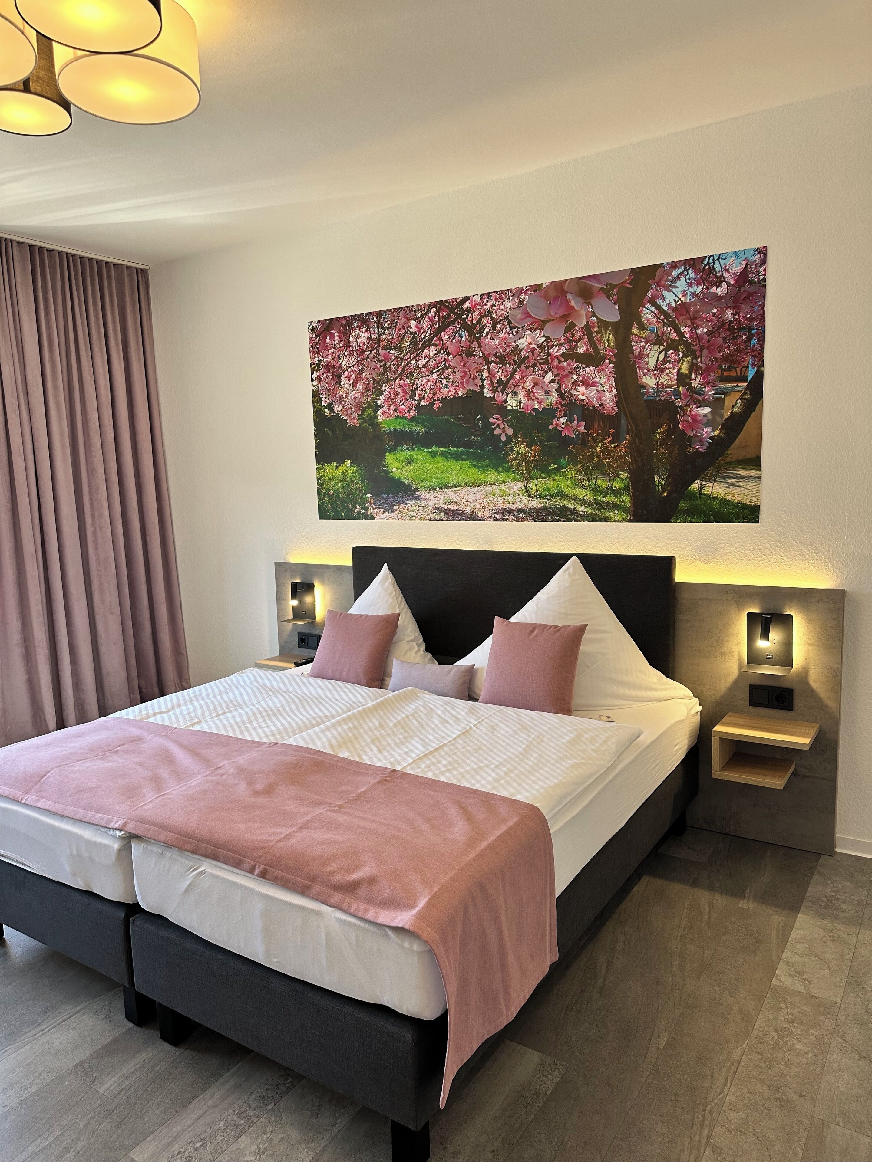 Ein Doppelbett mit Kopfteil, zwei Nachttische mit Lampen, ein großes Wandbild mit Kirschblüten und ein Fenster mit Vorhängen.