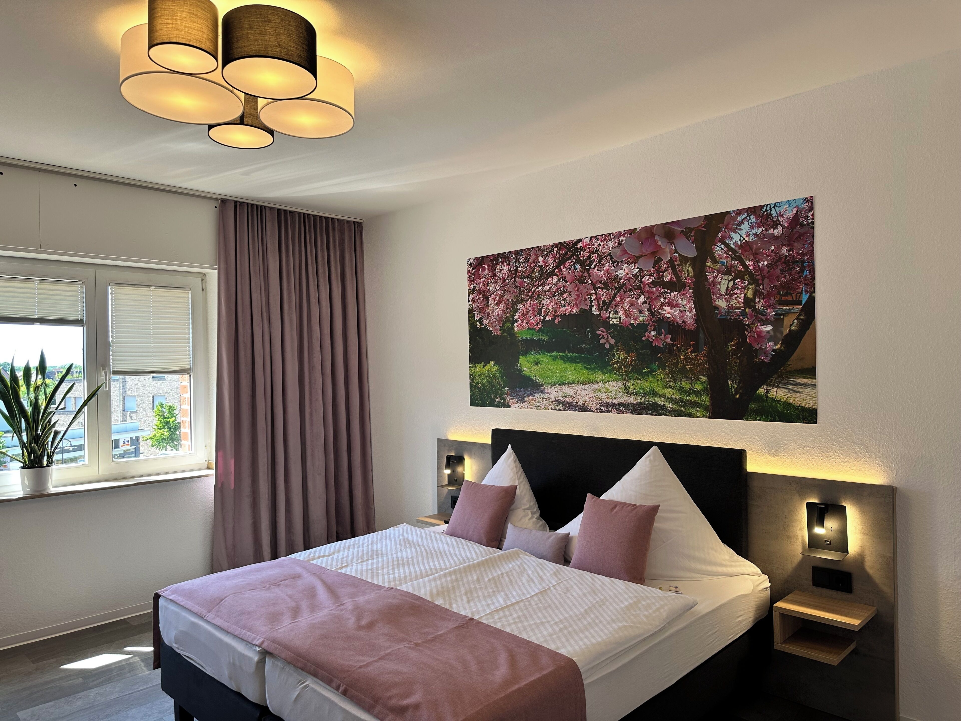 Une chambre à coucher avec un lit, une fenêtre avec des stores, une plante en pot, un tableau représentant des fleurs de cerisier et un luminaire fixé au mur.