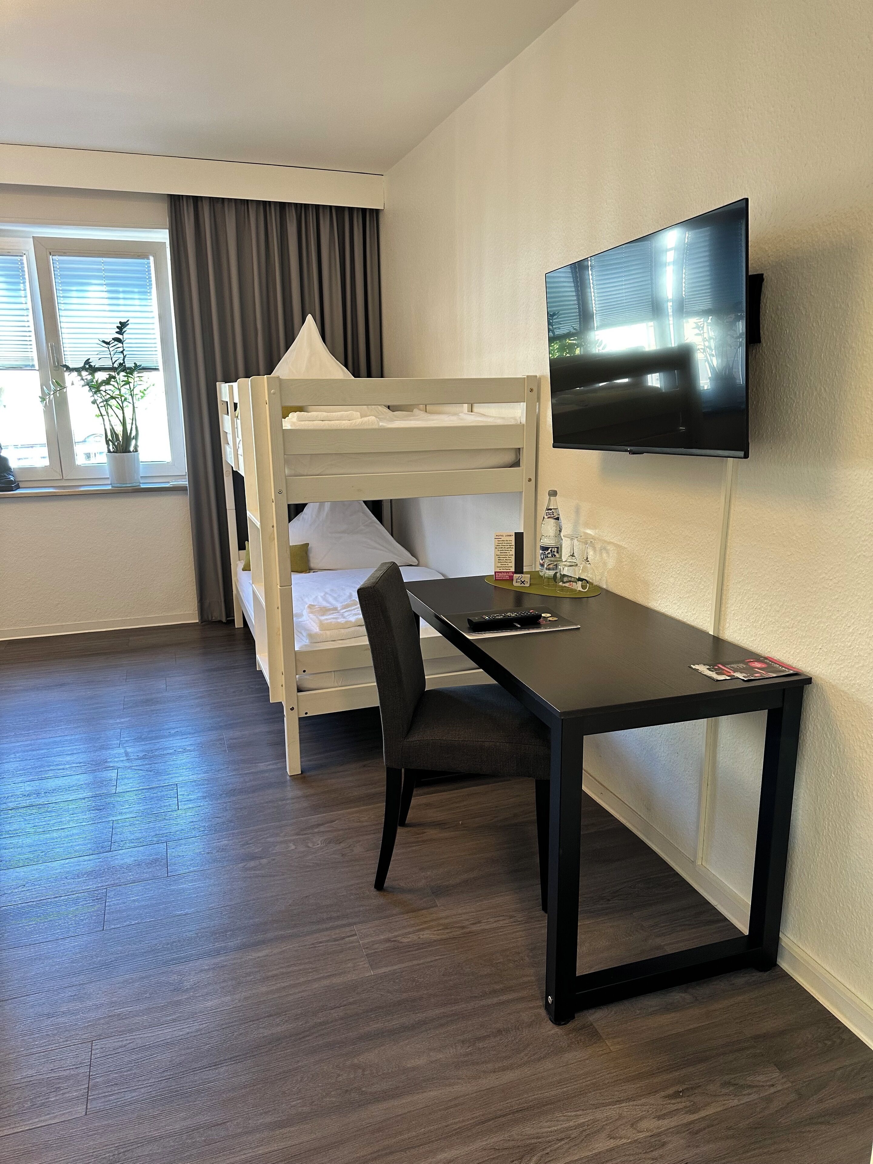 A hotel room with a bunk bed, a desk with a chair, a television, and a window with curtains.