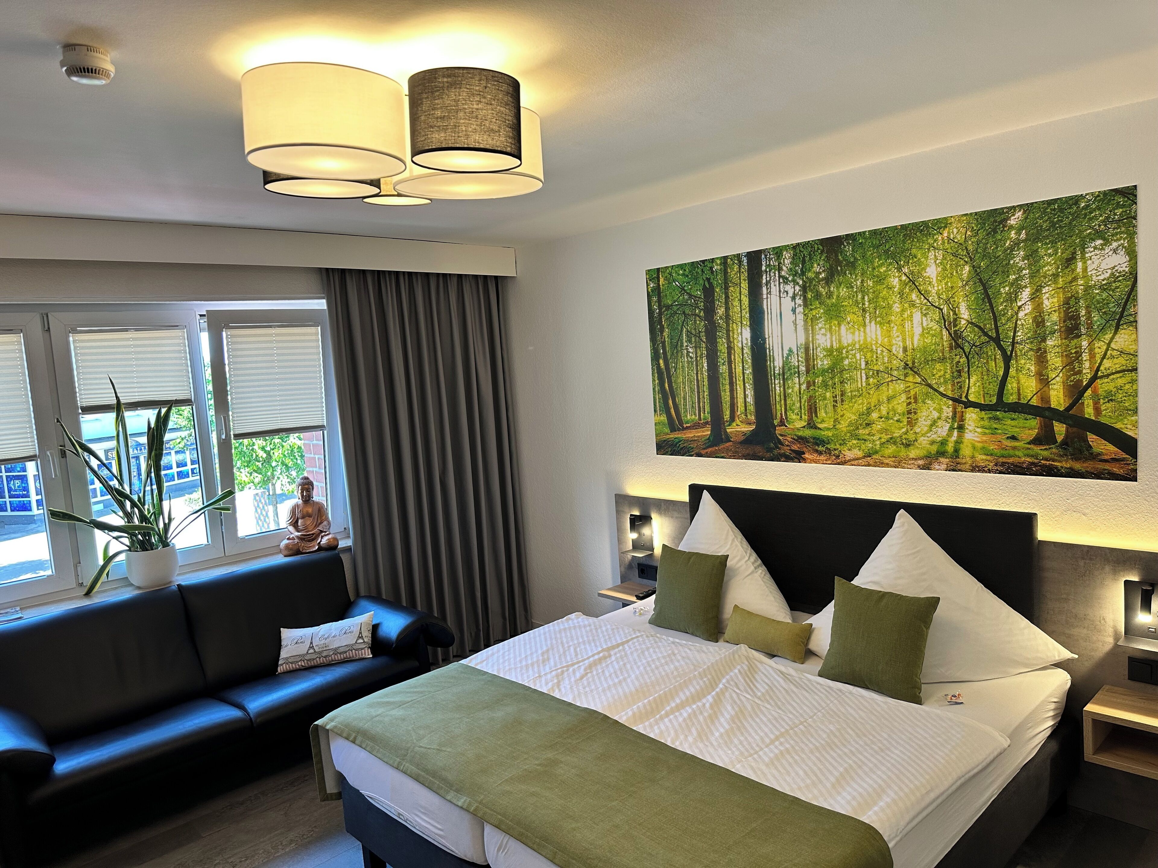 Een hotelkamer met een bed, een bank, een raam met gordijnen, een kamerplant en een schilderij met een boslandschap aan de muur.