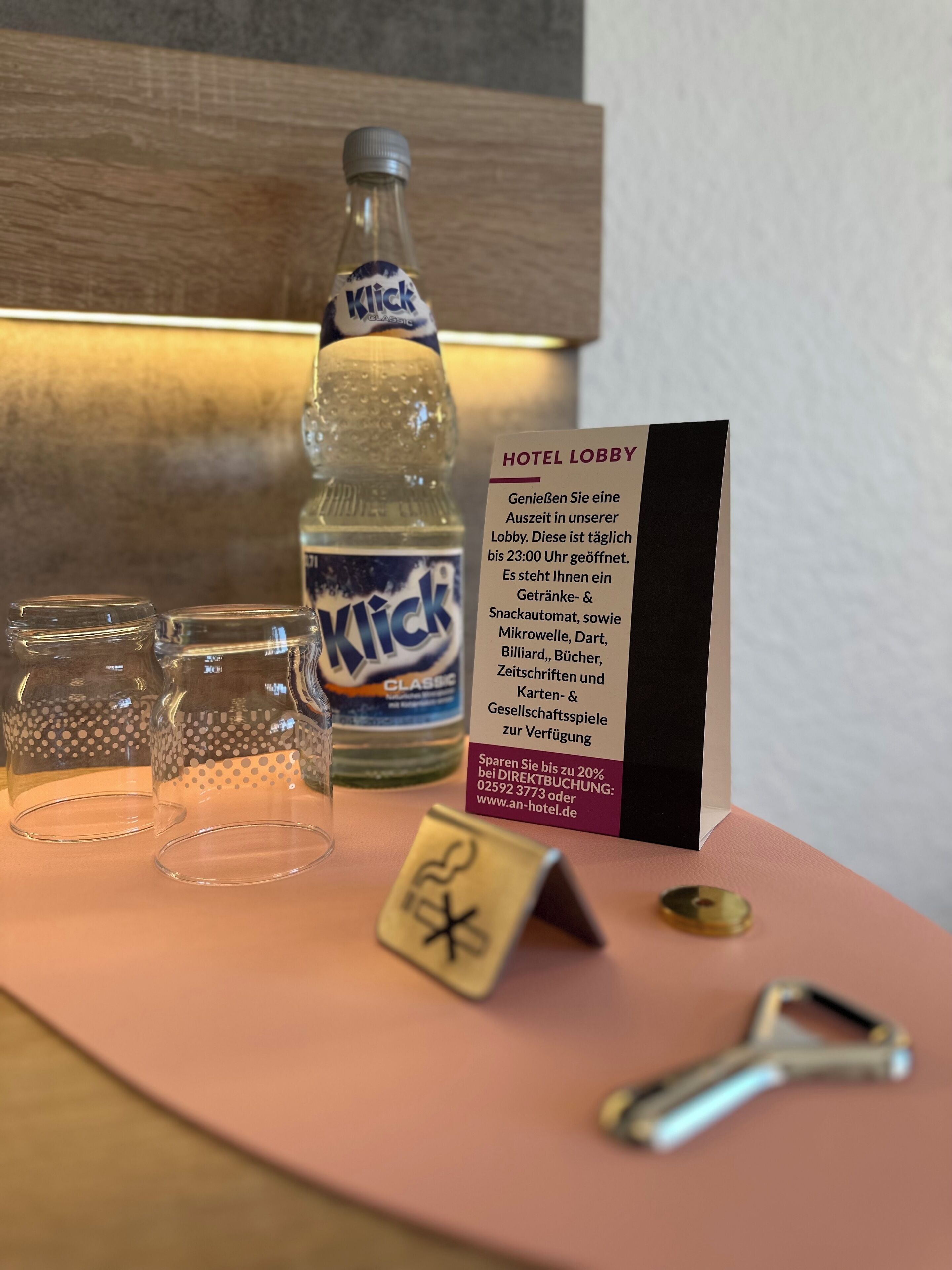 Un vestíbulo de hotel con una botella de agua KICK Classic, dos vasos vacíos, un cartel que detalla los servicios del vestíbulo y una llave.