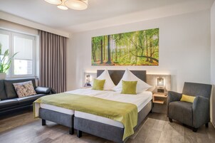 Un dormitorio moderno con una cama grande, un sillón gris, un sofá y un cuadro con motivos forestales.