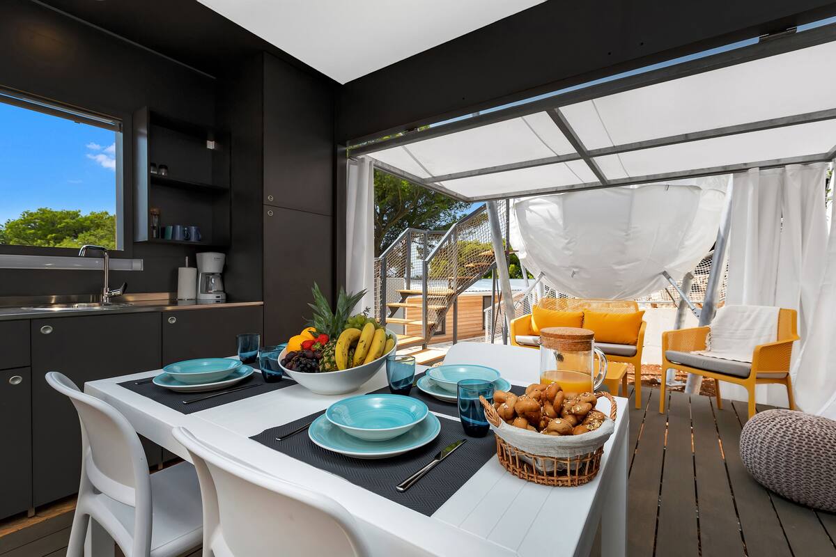 Une cuisine moderne avec un coin repas, où une table est dressée avec des assiettes, des verres et un panier de fruits.