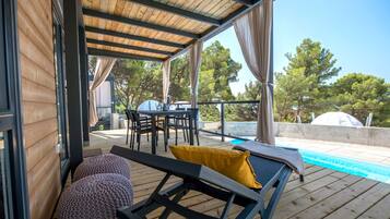 A covered outdoor area with a pool, wooden deck, lounge chairs, and a table with chairs.