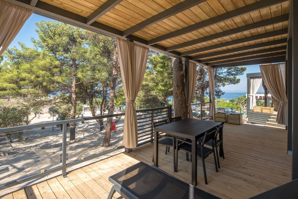 Une terrasse extérieure en bois, avec une table et des chaises, donnant sur une plage bordée d’arbres et offrant une vue dégagée sur la mer.