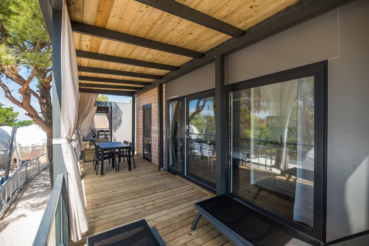 Un espace extérieur abrité doté d’une terrasse en bois, d’une table et de chaises, et de grandes portes vitrées donnant sur un patio.