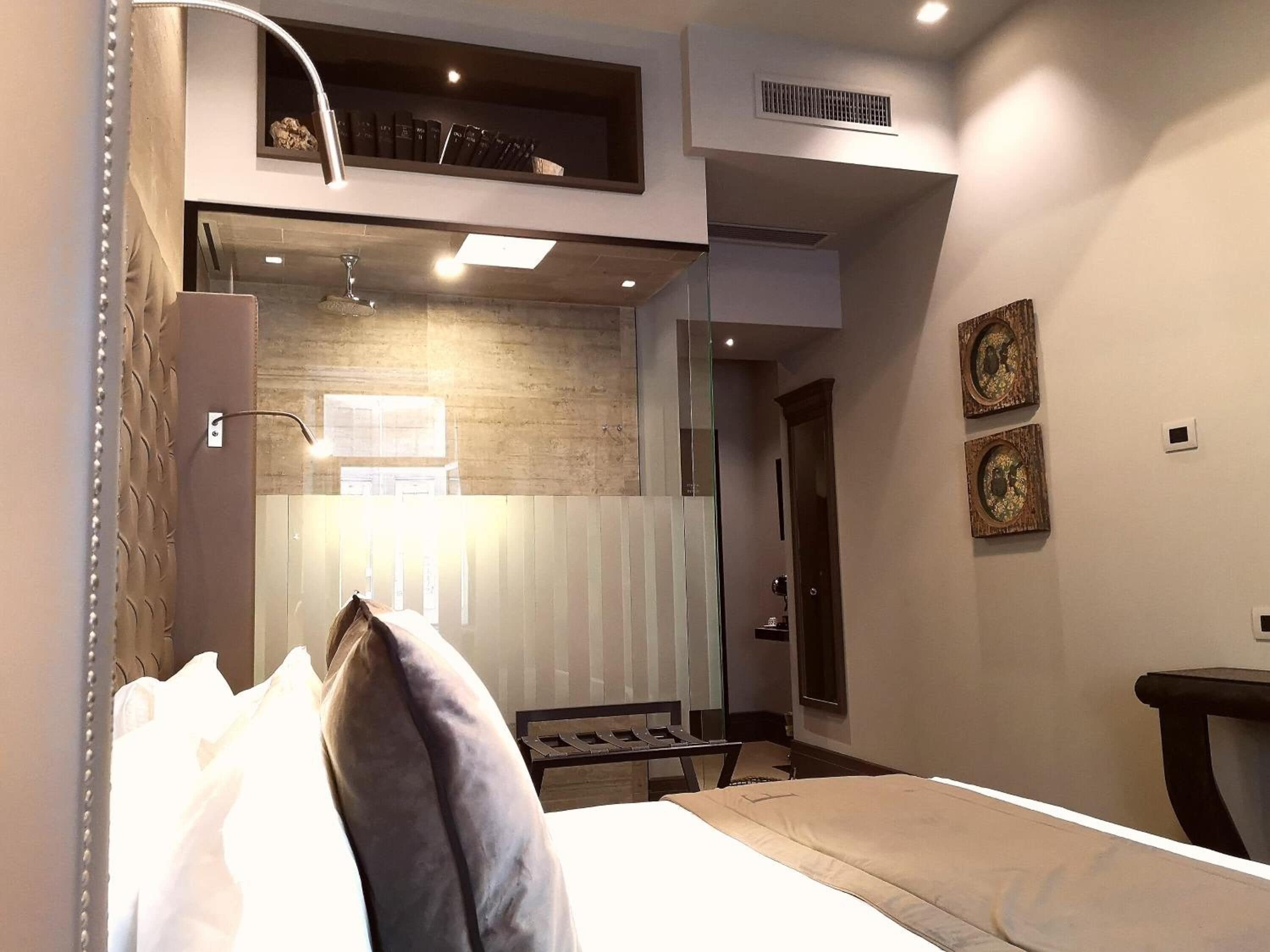 Habitación de hotel moderna con cama, escritorio y pequeño rincón de descanso. Se aprecia una ventana con persianas, un radiador y una estantería con libros.