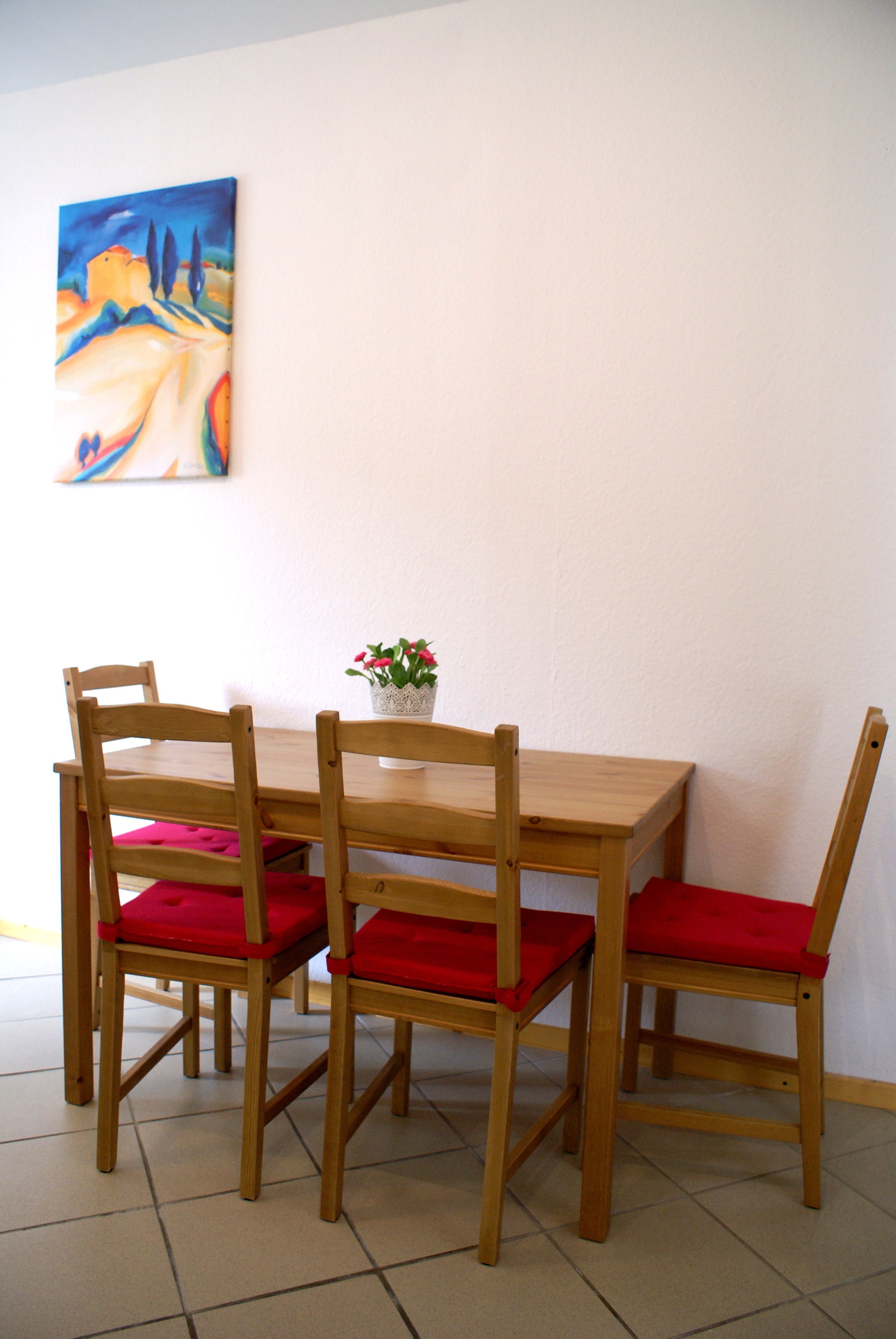 Un comedor con una mesa de madera y cuatro sillas, cada una con cojines rojos. En la mesa hay una pequeña planta en maceta con flores rosadas.