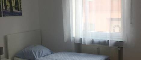A bedroom with two single beds, a nightstand, a radiator, and a window with sheer curtains.