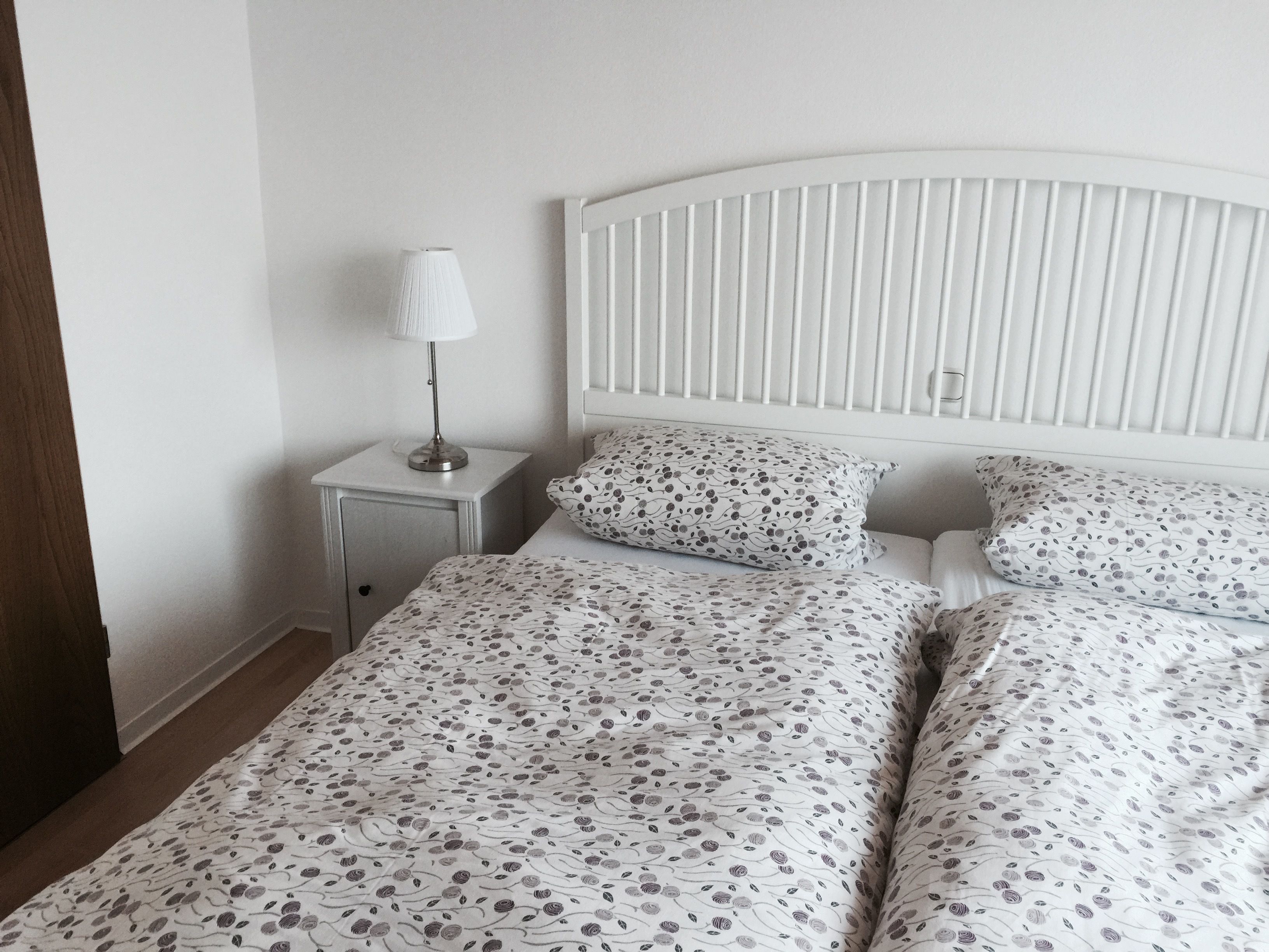 A bedroom with a white bed, a nightstand with a lamp, and a white headboard.