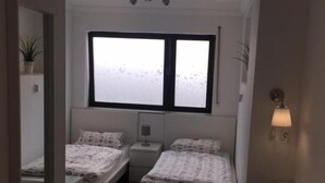 A small, well-lit bedroom with two single beds, a nightstand, a bedside table, a mirror, and a window with frosted glass. - Pension Lindenhof (Limburg an der Lahn)