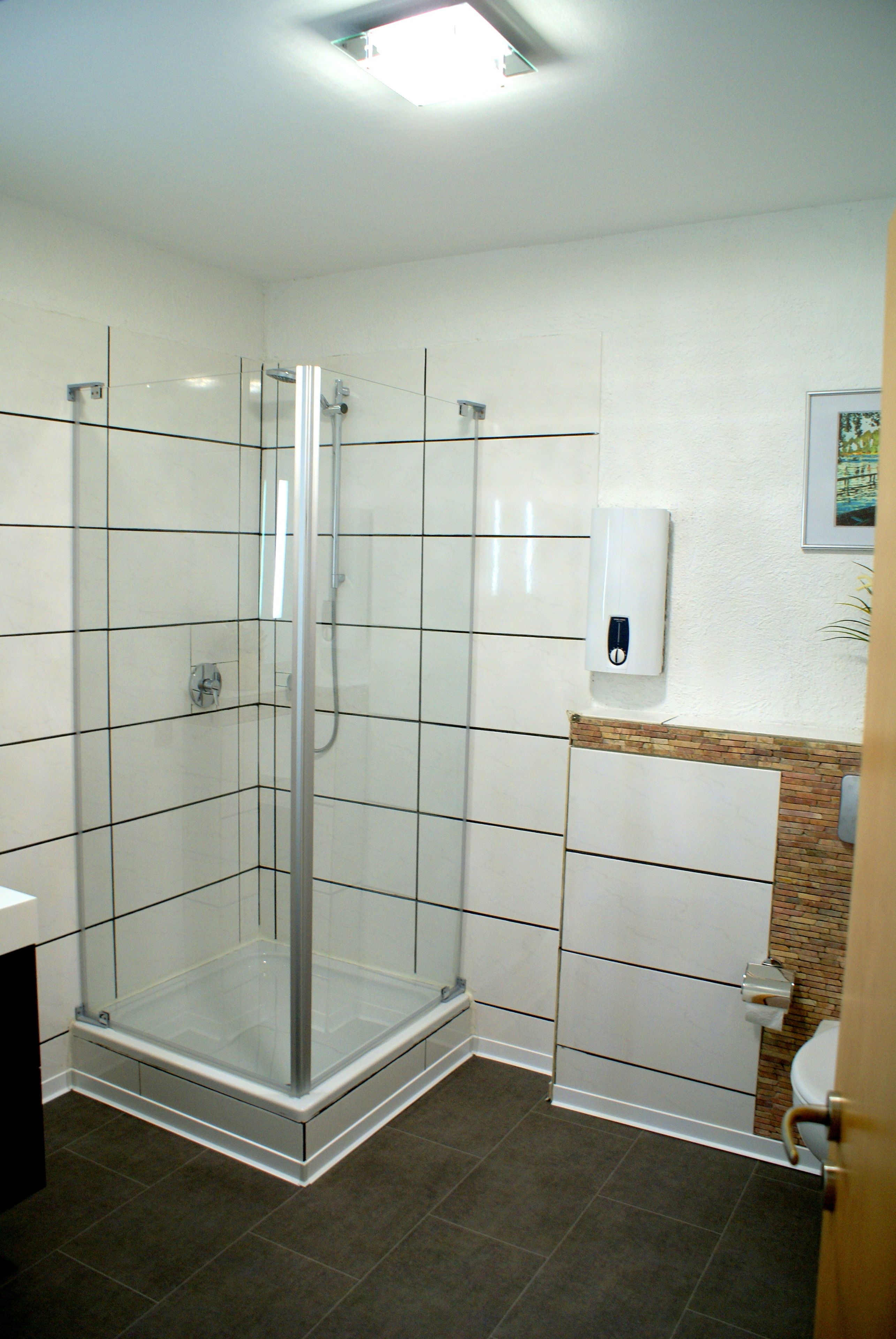 A modern bathroom with a glass-enclosed shower, white tiled walls, a toilet, and a hand sanitizer dispenser.