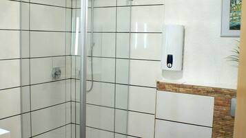 A modern bathroom with a glass-enclosed shower, white tiled walls, a toilet, and a hand sanitizer dispenser.
