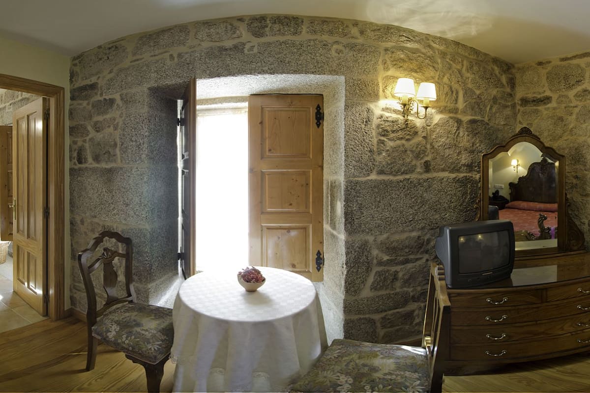 A bedroom with stone walls, a wooden bed, a dresser, a chair, and a table with a bowl.