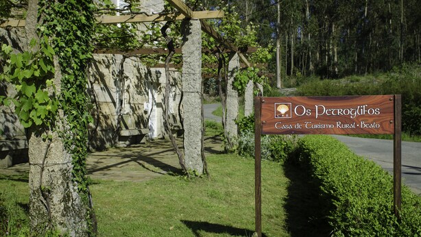 A rustic stone building with a wooden pergola, surrounded by greenery and a sign for "Os Petroglifos Casa de Turismo Rural".