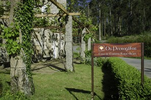 A rustic stone building with a wooden pergola, surrounded by greenery and a sign for "Os Petroglifos Casa de Turismo Rural". - Casa de Turismo Rural Os Petroglifos (Boiro)