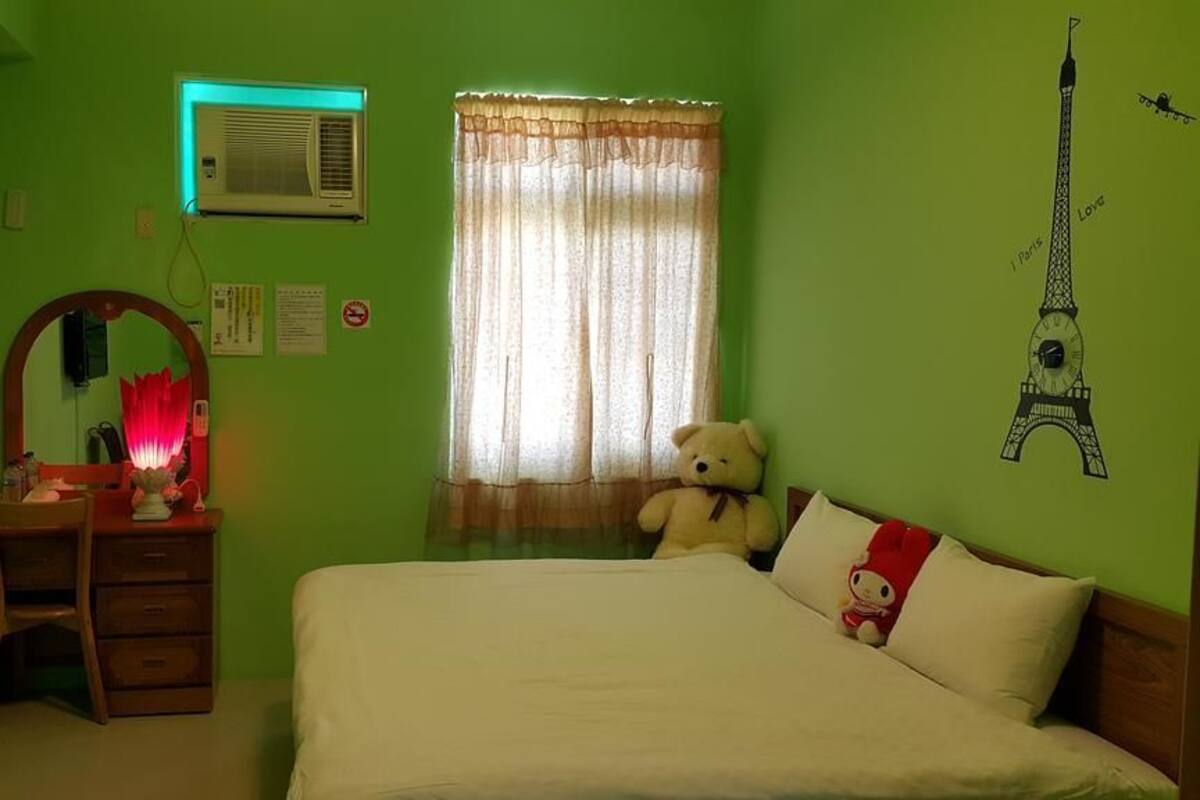 Una habitación con una cama, un escritorio, una silla, un peluche, un espejo, una ventana con cortinas y un dibujo de la Torre Eiffel en la pared.