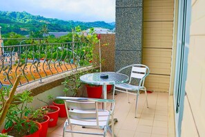 Un balcon avec une table ronde et deux chaises, des plantes en pot et une vue sur des collines verdoyantes.