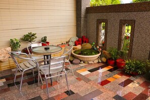 Un espace patio avec une table ronde et des chaises, un petit bassin avec des plantes, ainsi que des plantes en pot et des roches disposées dans cet espace.