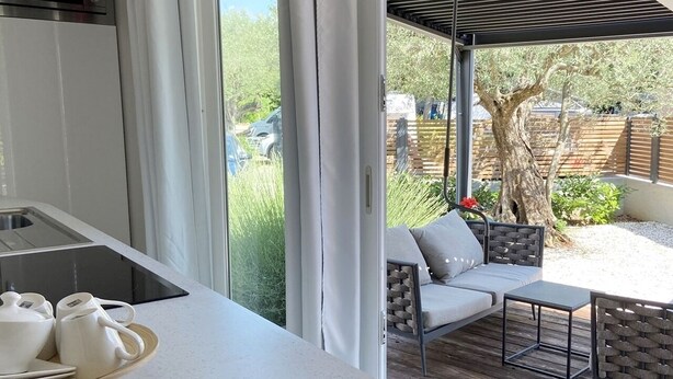 A modern kitchen with a large window, a dining area with a sofa and table, and a view of a garden and tree. - Ulika Rovinj Camping (Rovinj)