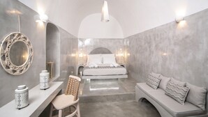 A modern bedroom with a bed, a sofa, a chair, and a vanity with a mirror and decorative items. - Thetis Cave Houses (Santorini)