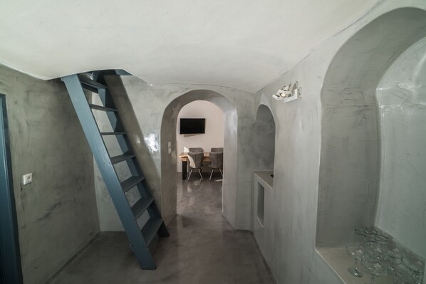 Un pasillo estrecho con una escalera metálica azul, un televisor de pantalla plana montado en la pared y una pequeña zona de estar con sillas y una mesa.