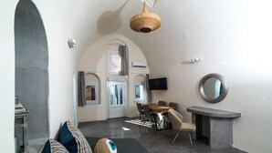 A modern interior with a dark sofa, a dining area with chairs, a TV, and a mirror. - Thetis Cave Houses (Santorini)