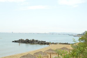 A beach with thatched umbrellas, a rocky breakwater, and a distant city skyline. - Alexandra Hotel (Sveti Vlas)