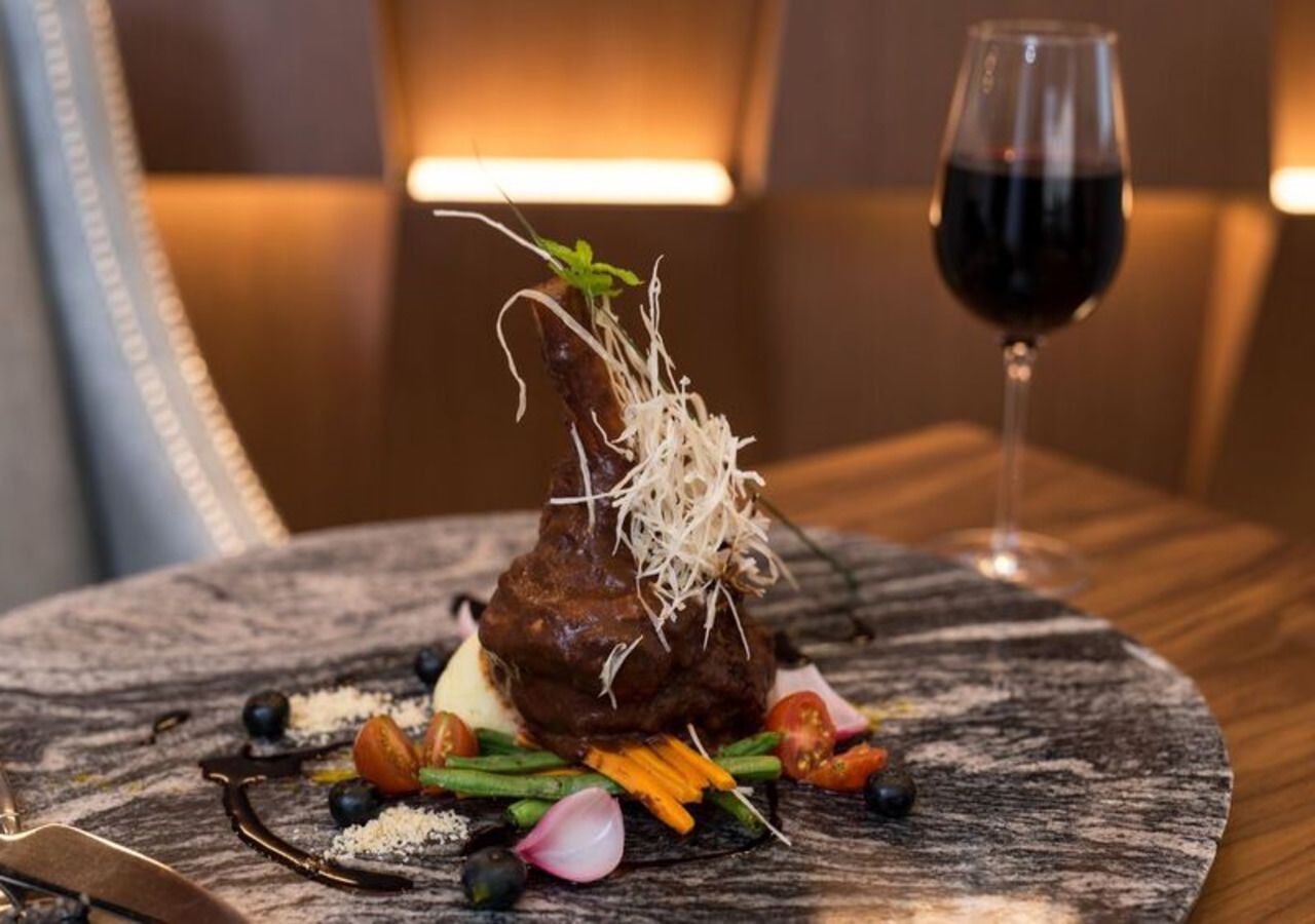 A plated dish with a steak, garnished with microgreens and vegetables, on a round table with a glass of red wine.