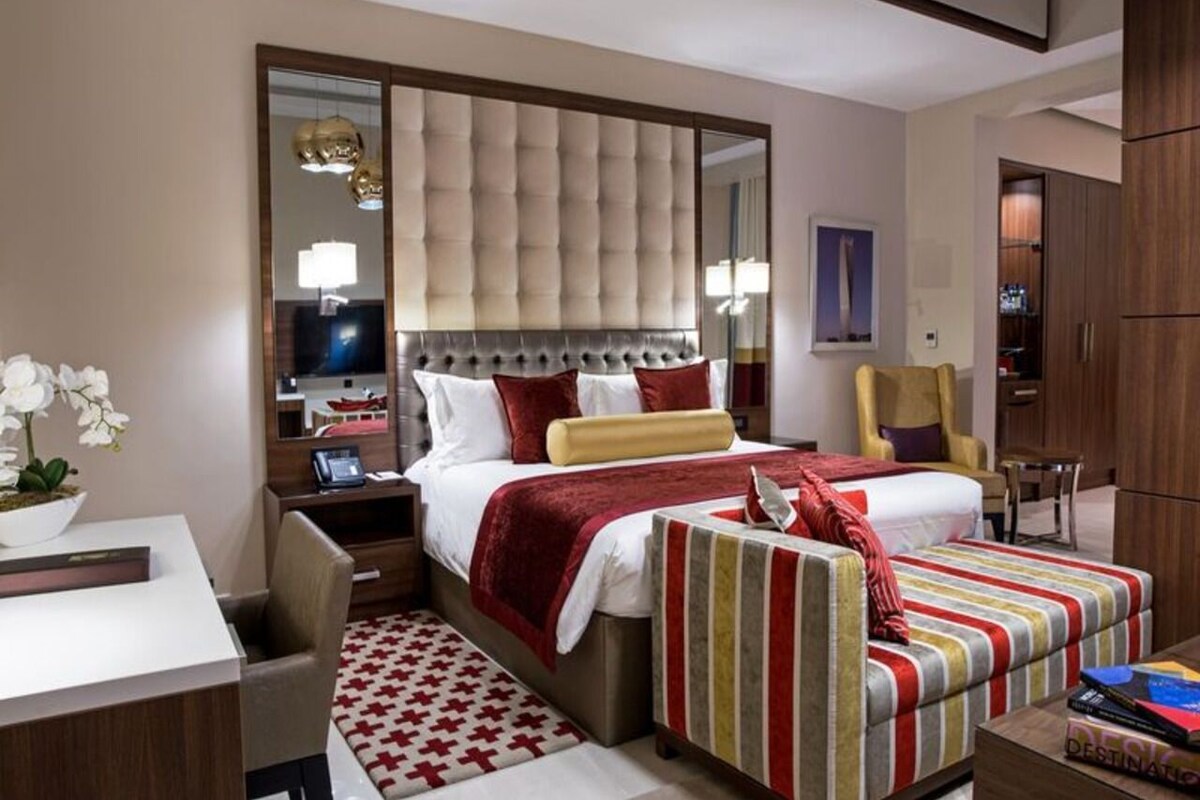 A hotel room with a large bed, a bedside table, a chair, a desk, and a mirror.
