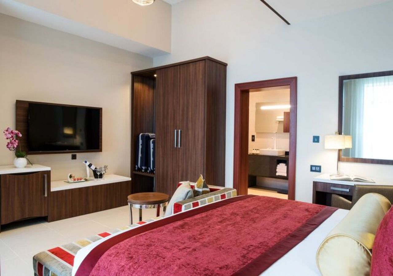 A modern hotel room with a large bed, a TV, a wardrobe, a desk, and a bathroom visible through an open door.