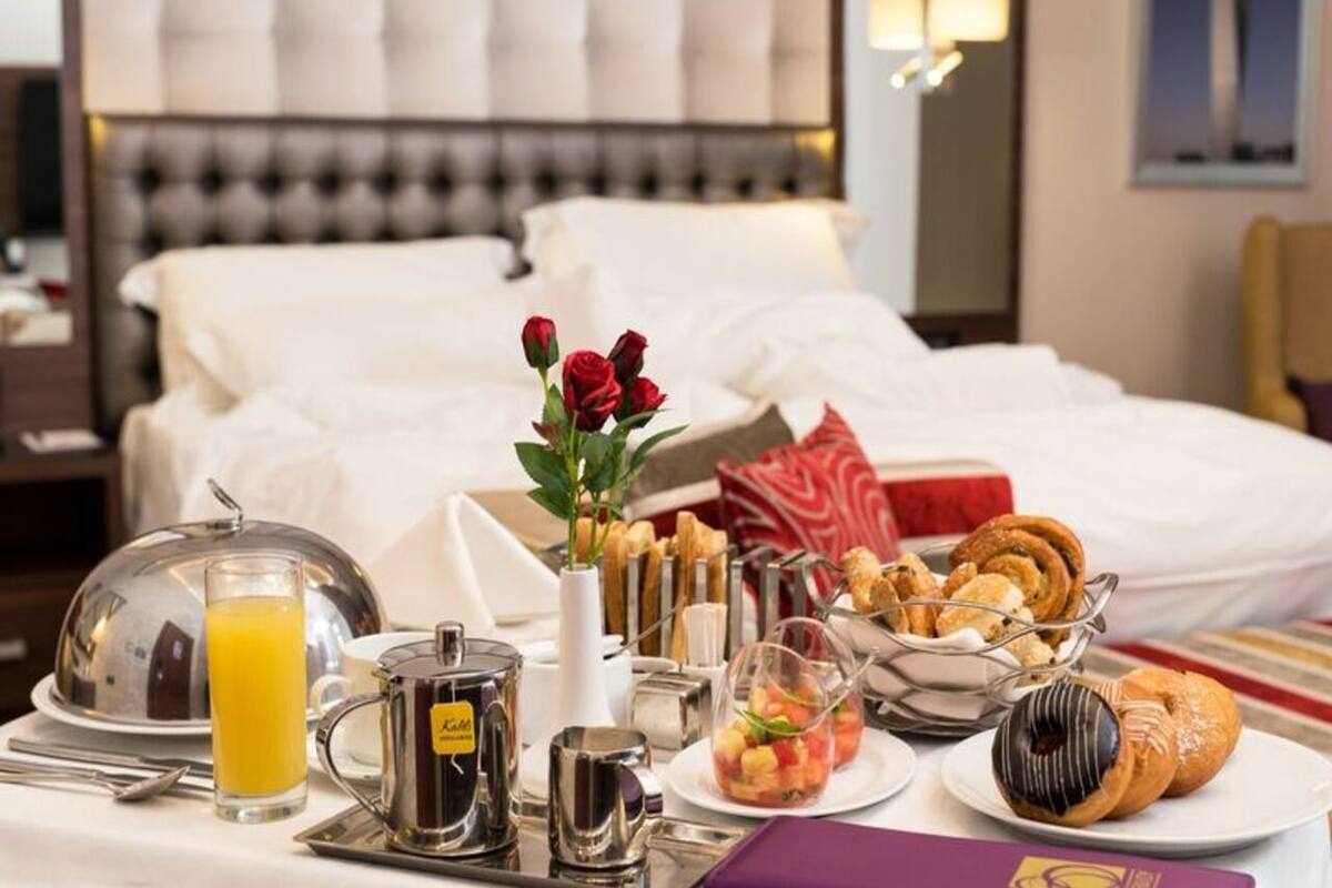 A hotel room with a neatly made breakfast table, a bed with white linens, and a decorative vase with red flowers.