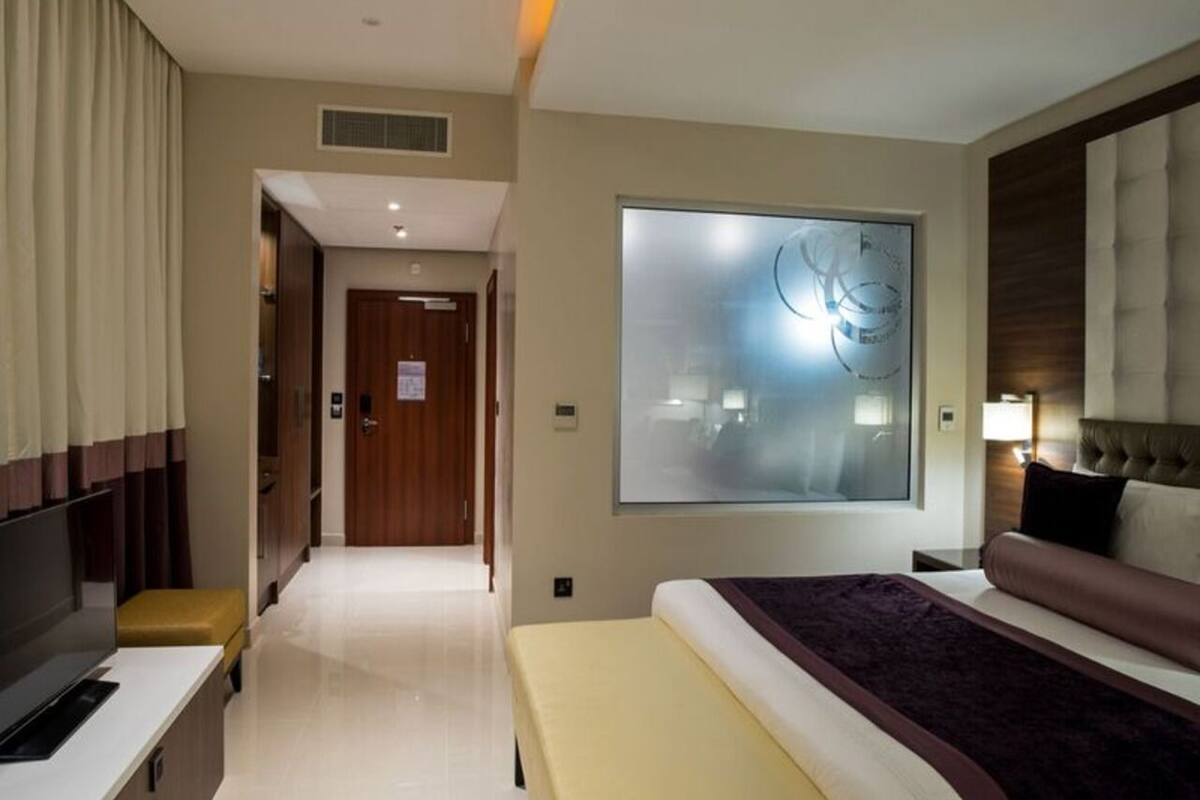 A hotel room with a bed, a desk, a chair, a large window, and a door leading to another room.