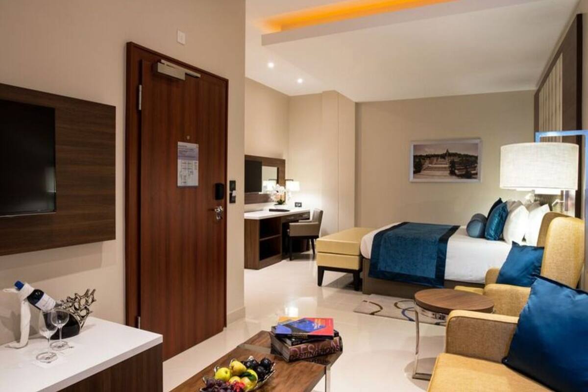 A modern hotel room with a flat-screen TV, a desk with a chair, a bed with blue bedding, a sofa, and a coffee table with books and fruit.