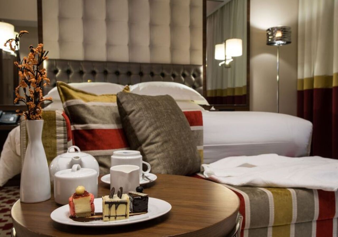 A hotel room with a bed, a bedside table with a vase, teapot, cups, and a plate of cake.