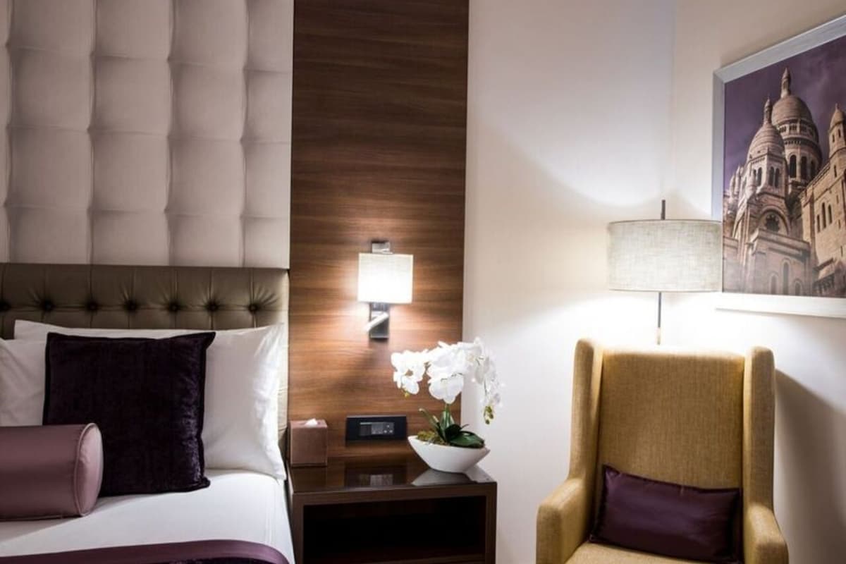 Una habitación de hotel con cabecera acolchada, una mesita de noche con lámpara, una silla y un aplique de pared.