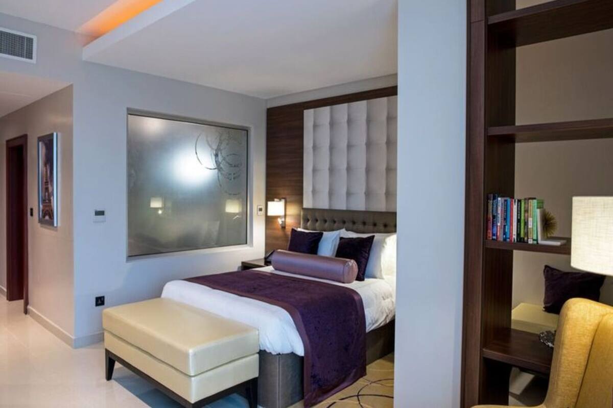 A modern hotel room with a large bed, a seating area, a bookshelf, and a large window.
