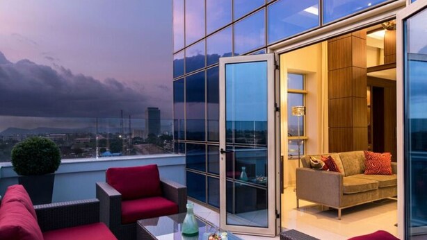 A modern outdoor seating area with wicker furniture and red cushions, a glass door leading to an interior space with a sofa and cushions, and a cityscape view in the background. - The Envoy Hotel (Abuja)