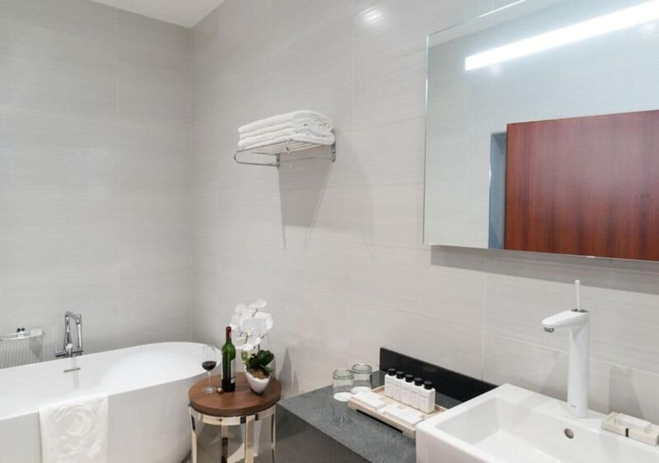 A modern bathroom with a bathtub, a sink, and a vanity. There is a towel rack on the wall and a small table with a plant and a bottle of wine.