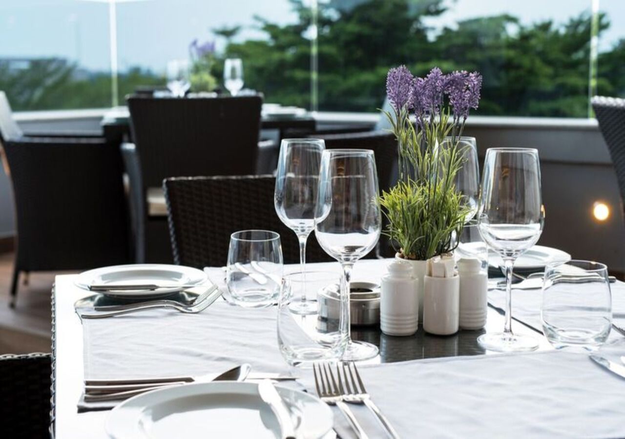 A dining table set for two with white tablecloths, clear glassware, and a small potted plant with purple flowers.