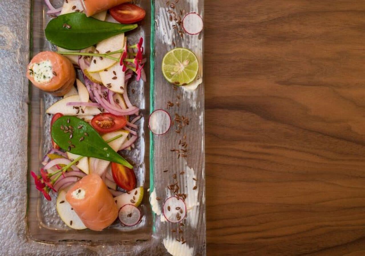 A wooden table with a glass dish containing a salad with tomatoes, onions, radishes, and a slice of lime.