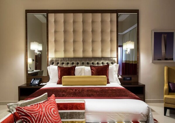 A hotel room with a large bed, a tufted headboard, a large mirror, a bedside table with a lamp, and a chair.