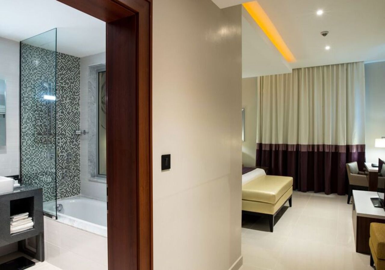 A modern hotel room with a glass shower, a bed, a desk, and a chair.