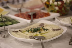 A buffet table with various dishes including a white creamy dish garnished with herbs and chickpeas, a red tomato-based dish, and a green salad.