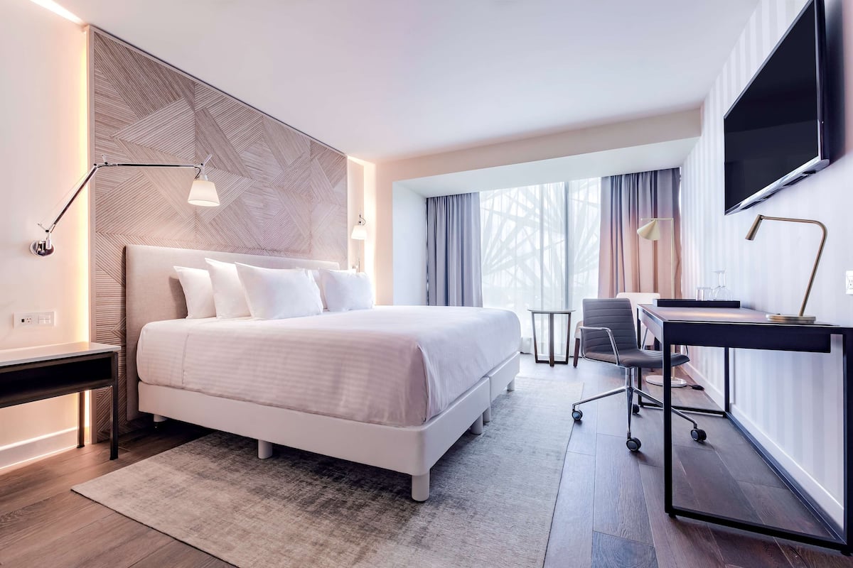 A modern hotel room with a large bed, a desk, a chair, a TV, and a window with curtains.