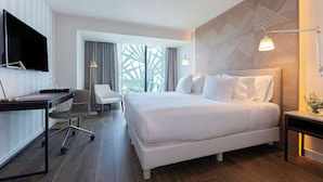 A modern hotel room with a large bed, a desk with a chair, a TV, and a view of greenery through the window. - NH Collection Merida Paseo Montejo (Mérida)