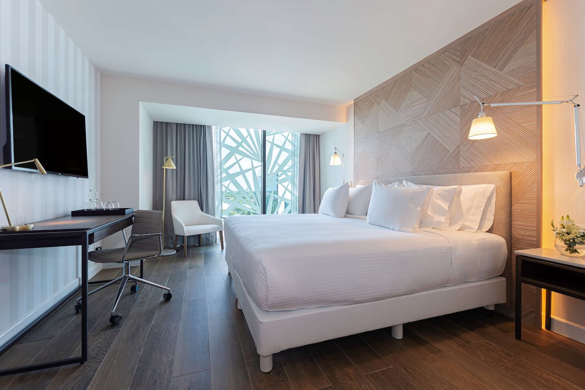 A modern hotel room with a large bed, a desk with a chair, a TV, and a view of greenery through the window.